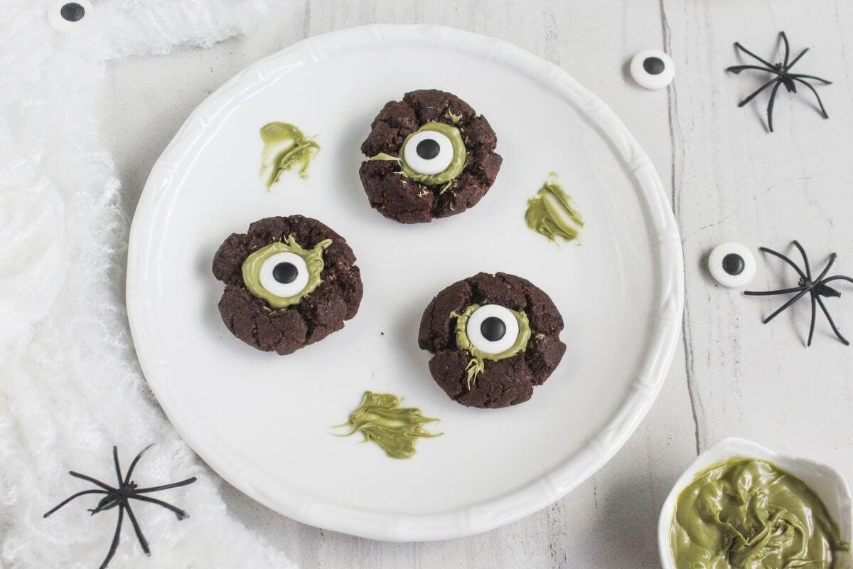 Three chocolate cookies with green frosting and candy eyes on a white plate, surrounded by plastic spiders and green frosting.