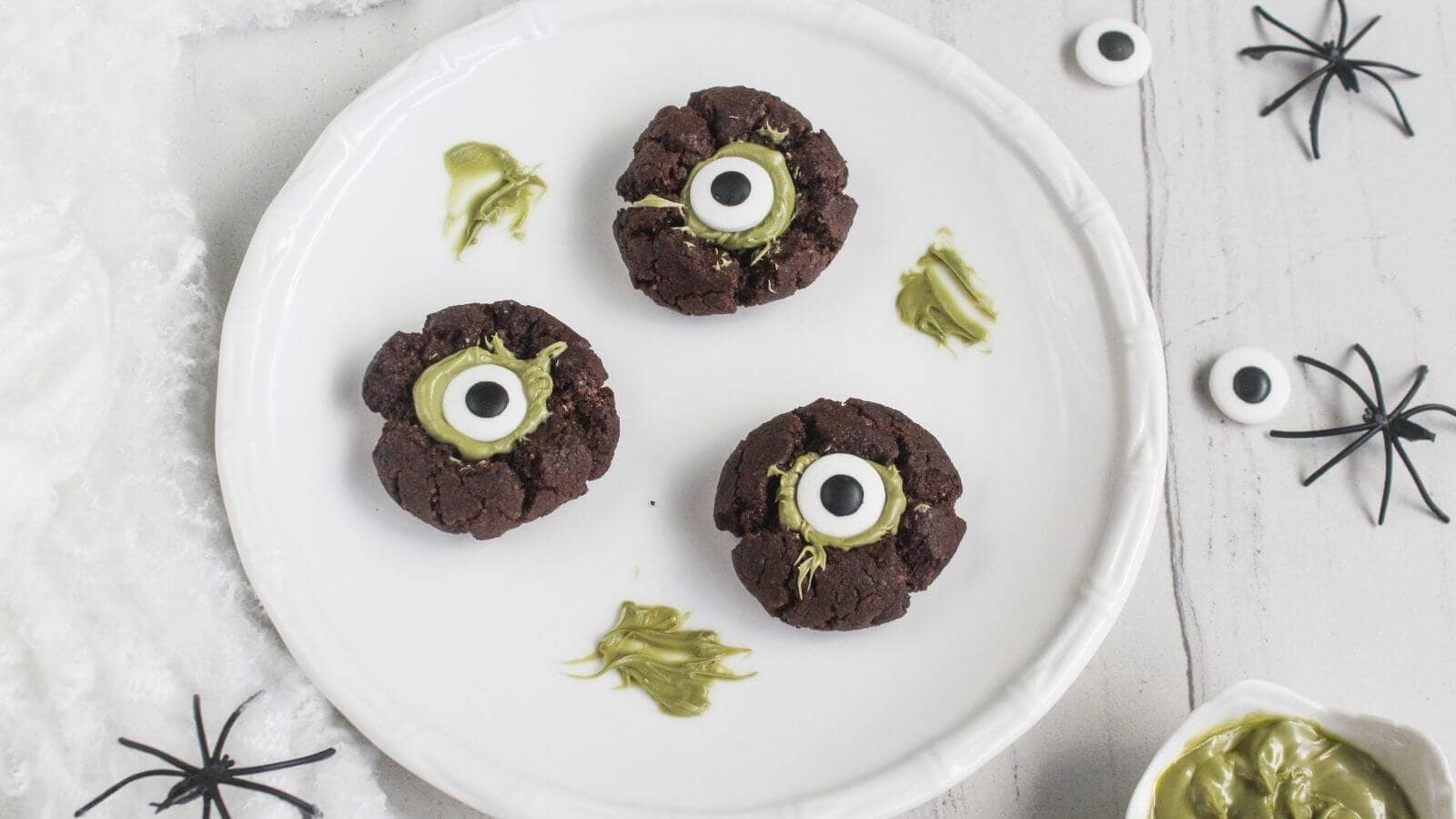 Three chocolate cookies with green frosting and candy eyes are arranged on a white plate, with plastic spiders and green icing on the side.