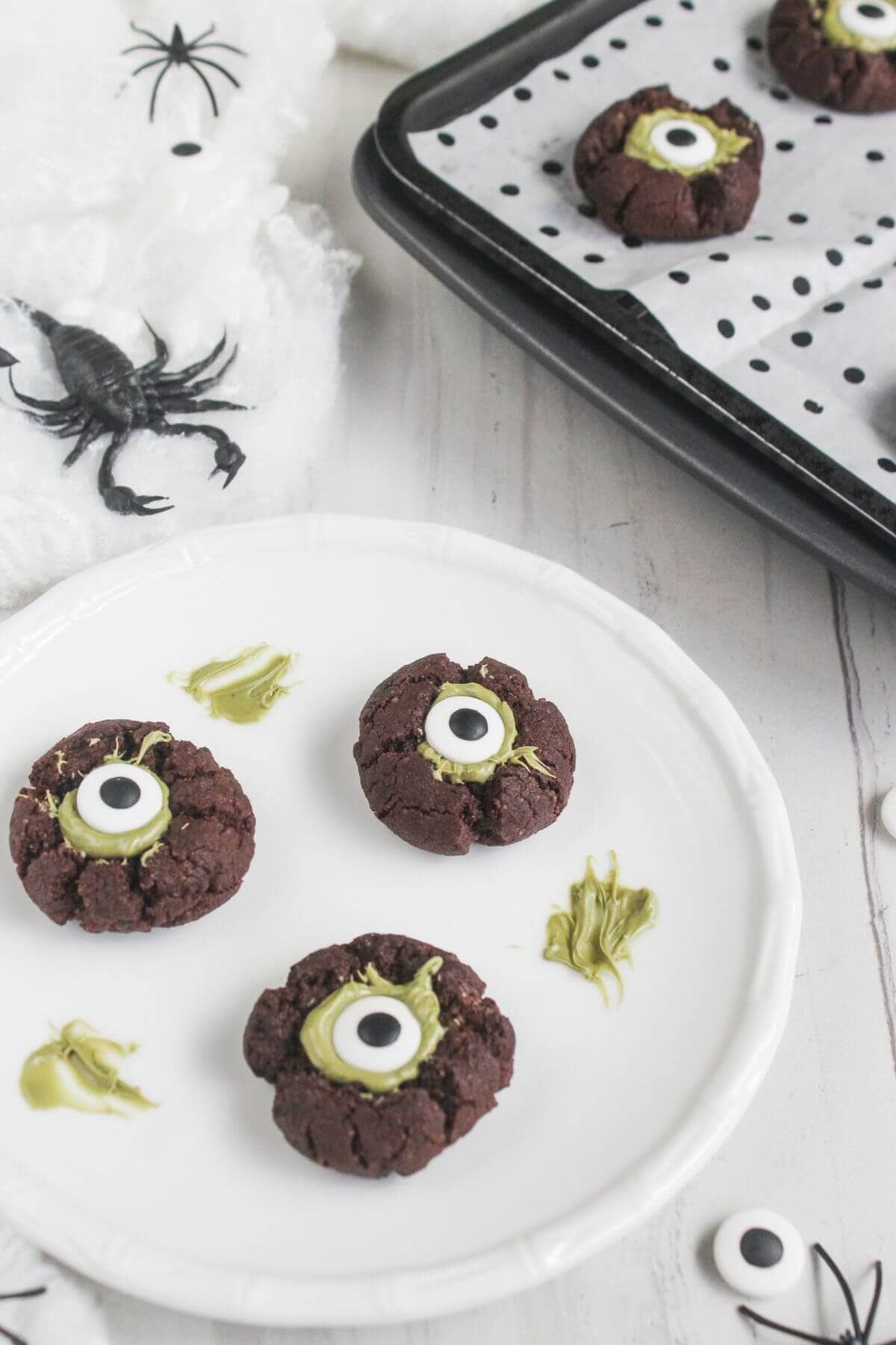 Three chocolate cookies with candy eyes and green icing on a white plate, next to a tray of more cookies and Halloween decorations like fake spiders and webbing.