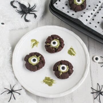 Three chocolate cookies decorated with green icing and candy eyeballs on a white plate, surrounded by fake spiders and a baking tray.