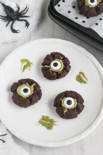Three chocolate cookies decorated with green icing and candy eyeballs on a white plate, surrounded by fake spiders and a baking tray.