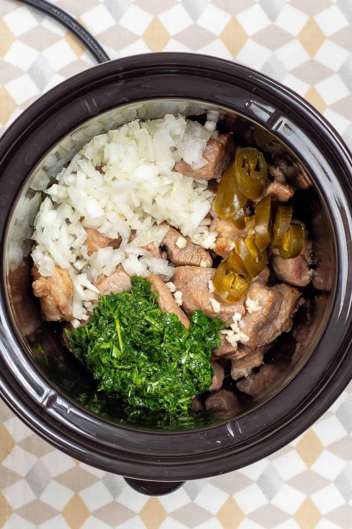 A slow cooker filled with raw meat chunks topped with diced onions, sliced jalapeños, and chopped cilantro.