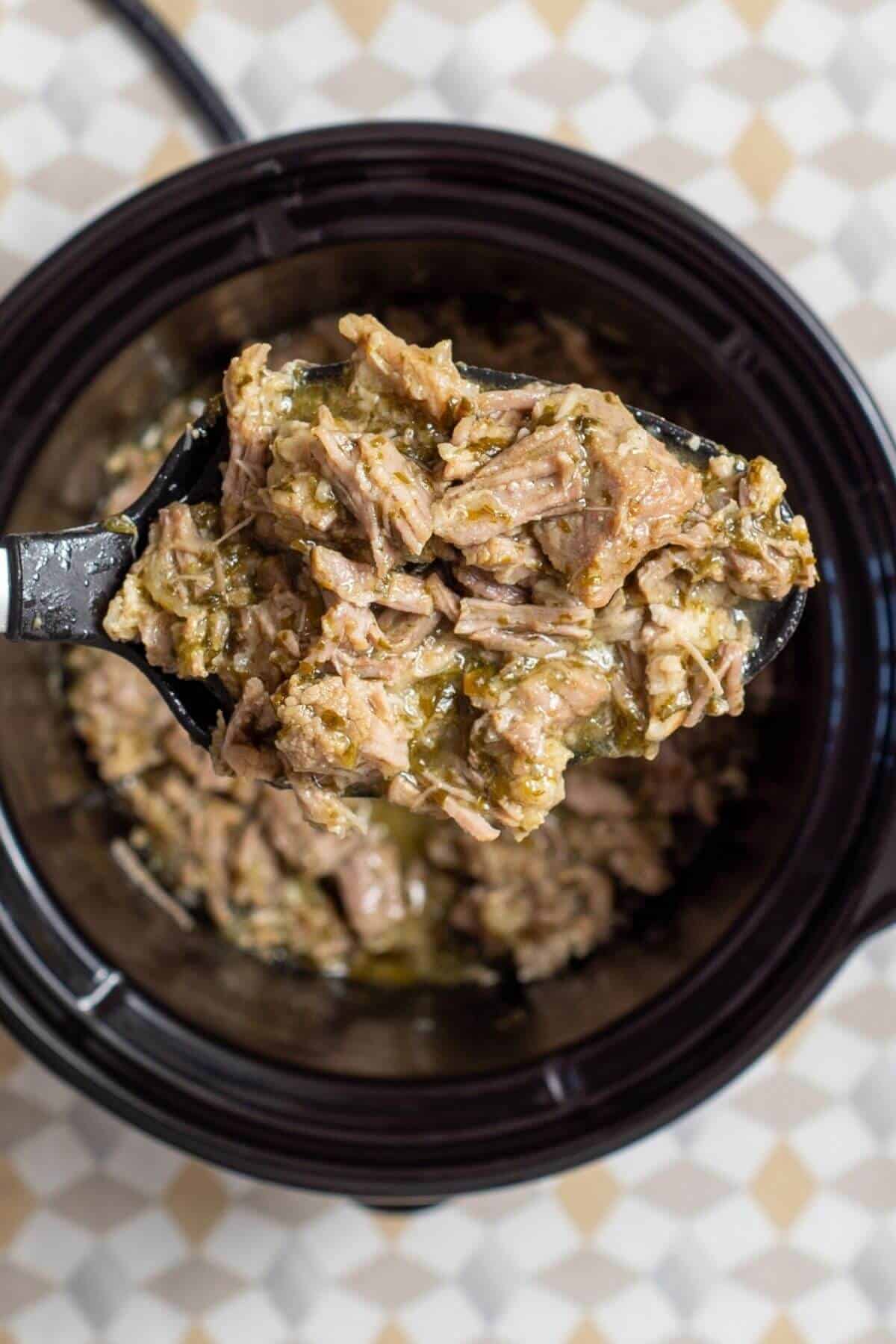 A black serving spoon lifting shredded pork in green sauce from a slow cooker.