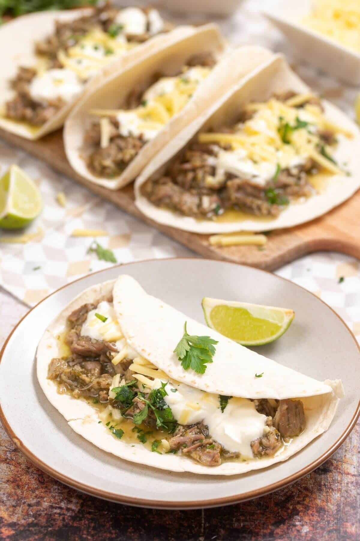 Three soft flour tacos filled with shredded meat, cheese, and sour cream on a plate and wooden board, garnished with cilantro and lime wedges.