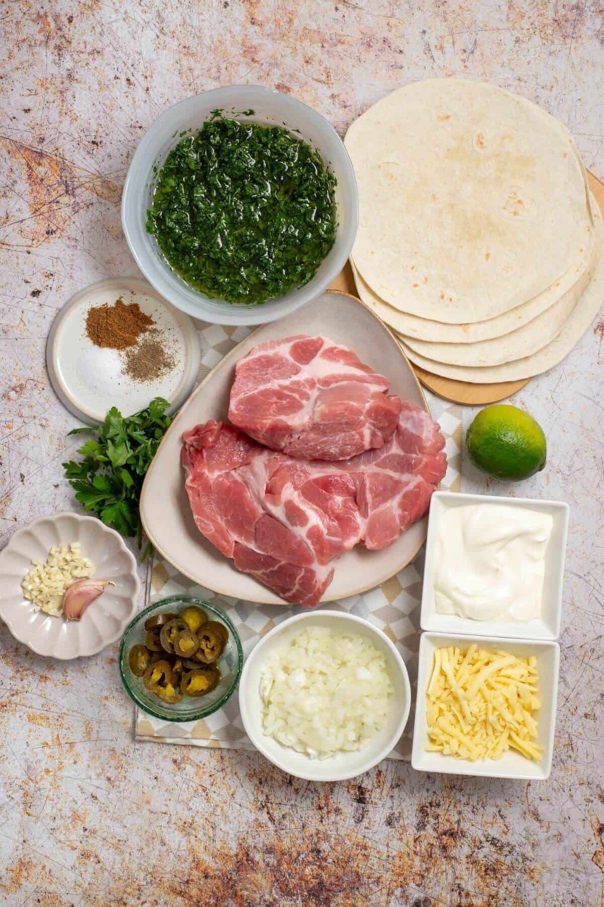 Raw pork shoulder, flour tortillas, a bowl of green herb sauce, a lime, sour cream, shredded cheese, chopped onion, sliced jalapeños, garlic, and spices arranged on a table.