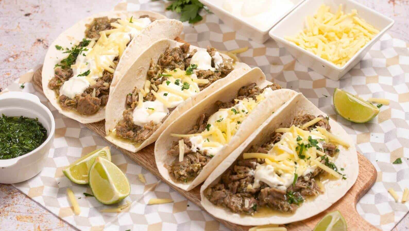 Four flour tortilla tacos filled with meat, sour cream, shredded cheese, and herbs, served on a wooden board with lime wedges and bowls of toppings.