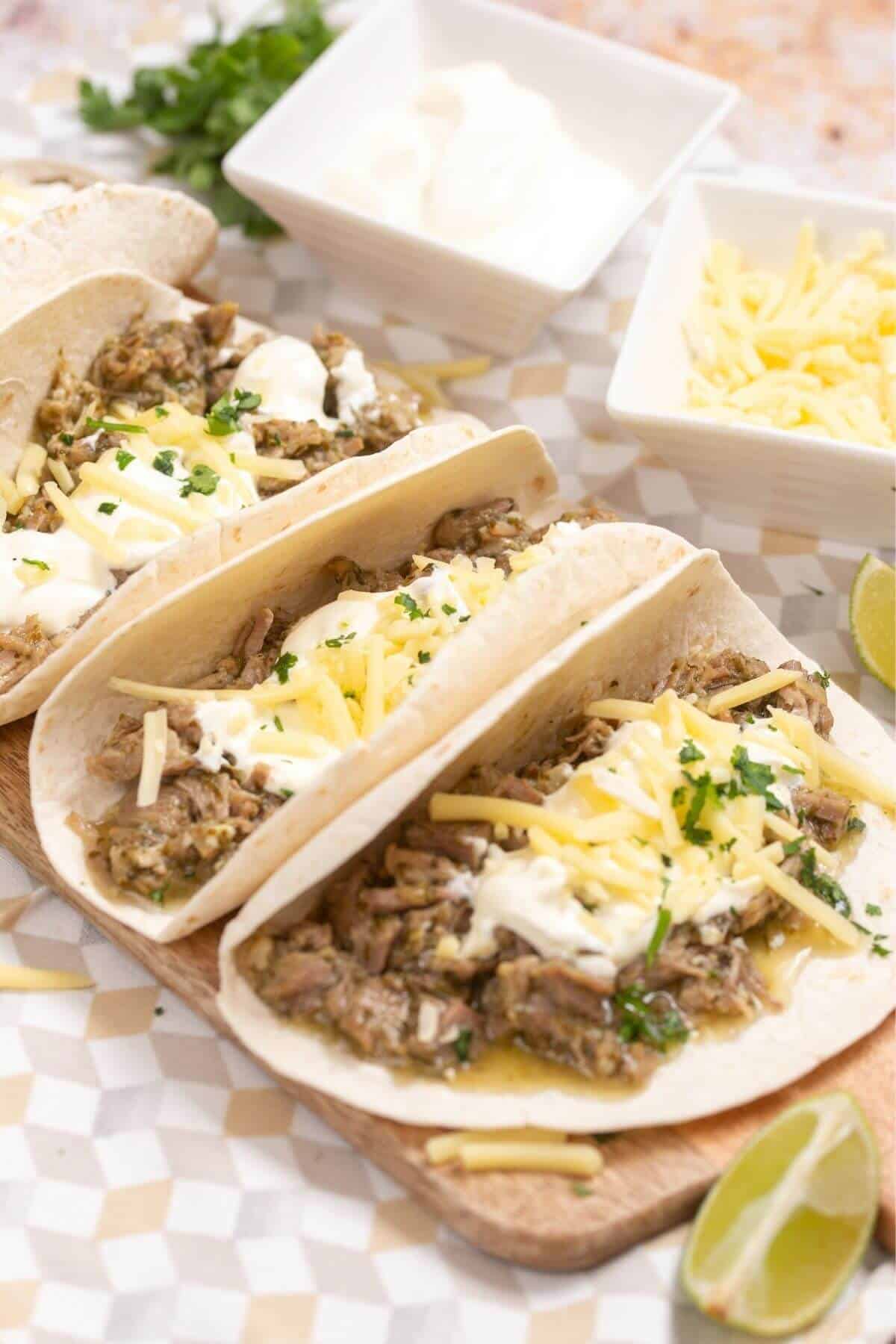 Three carnitas tacos on flour tortillas topped with shredded cheese, sour cream, and cilantro, served with sides of sour cream and cheese.