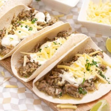 Three flour tortillas filled with shredded meat, sour cream, shredded cheese, and herbs, served on a wooden board.