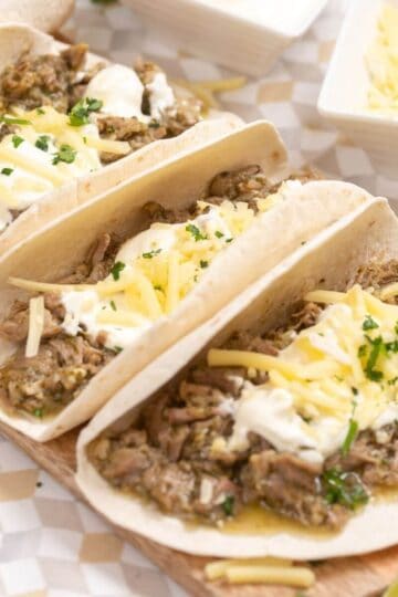 Three flour tortillas filled with shredded meat, sour cream, shredded cheese, and herbs, served on a wooden board.