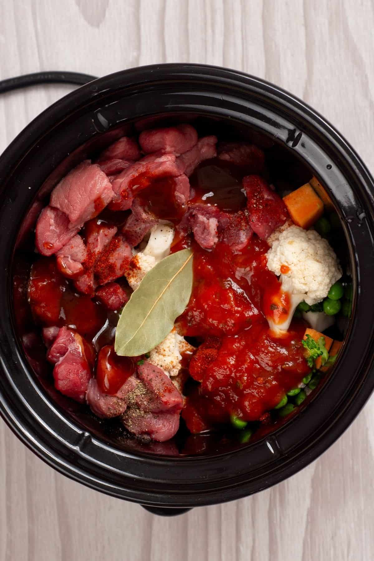 A slow cooker filled with raw beef chunks, cauliflower florets, peas, carrots, tomato sauce, and a bay leaf.
