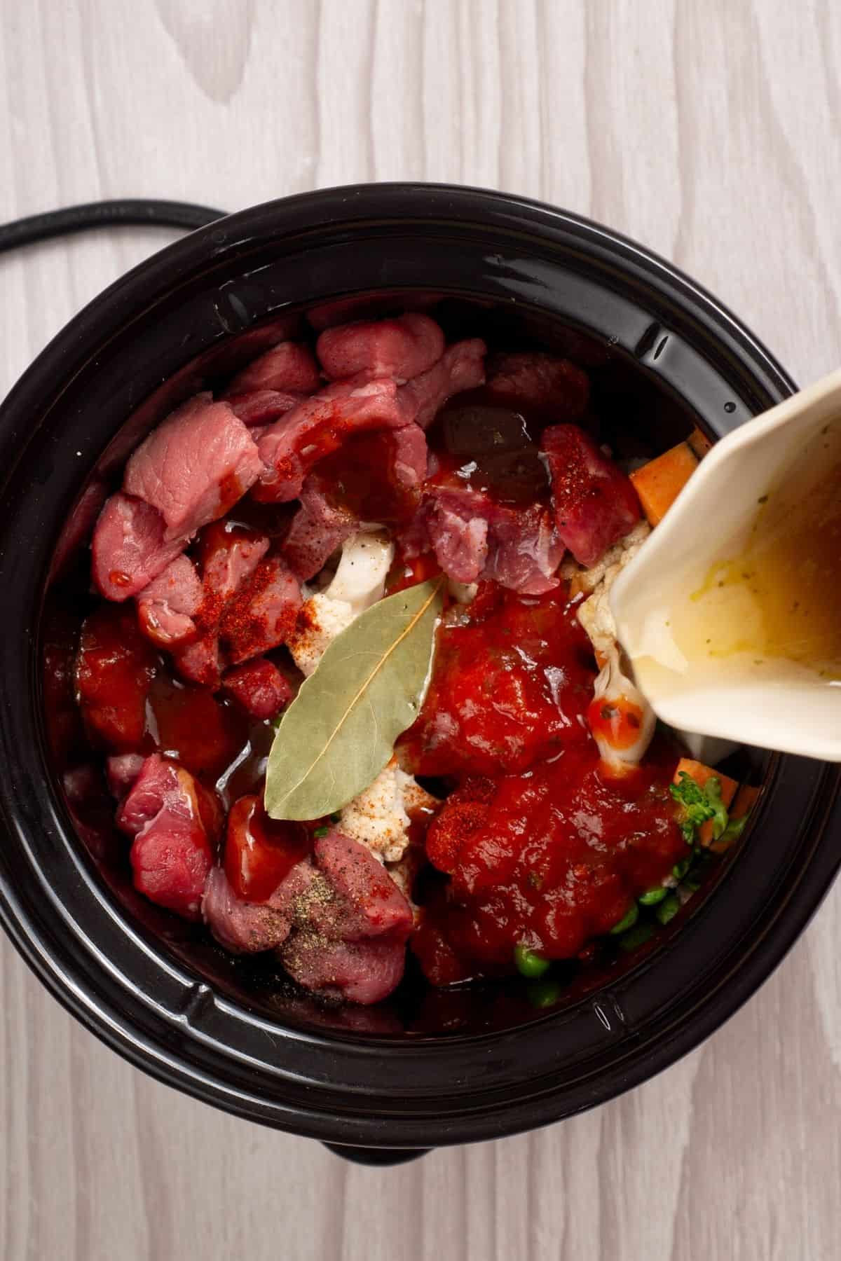 A slow cooker containing raw beef, vegetables, tomato sauce, and a bay leaf, with liquid being poured in from a pitcher.