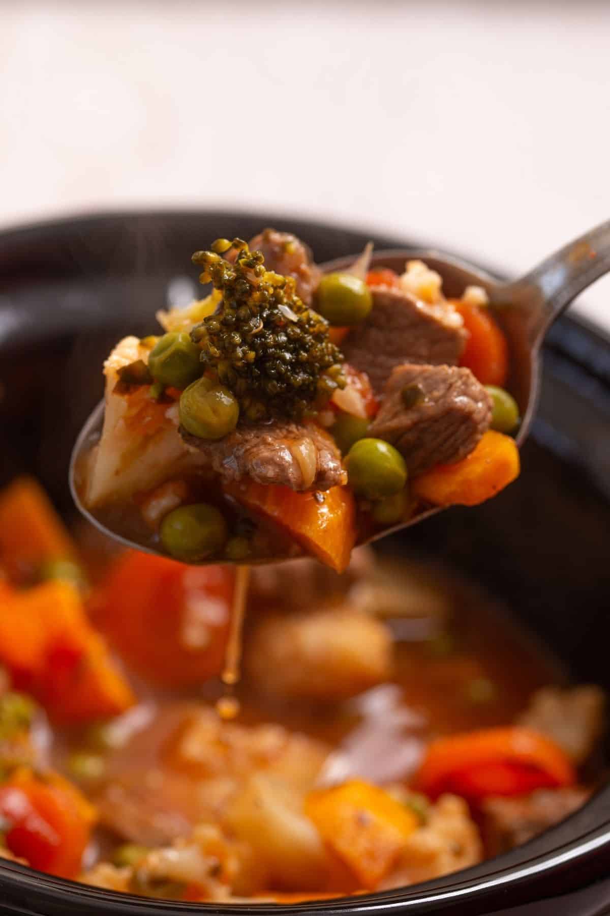 A metal spoon lifting a serving of beef and vegetable stew containing broccoli, peas, carrots, and cauliflower from a black bowl.
