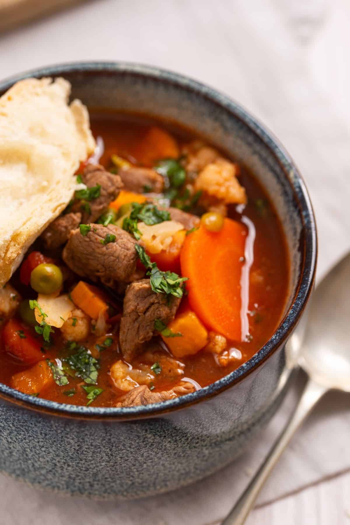 A bowl of beef stew with carrots and peas, served with a piece of crusty bread.