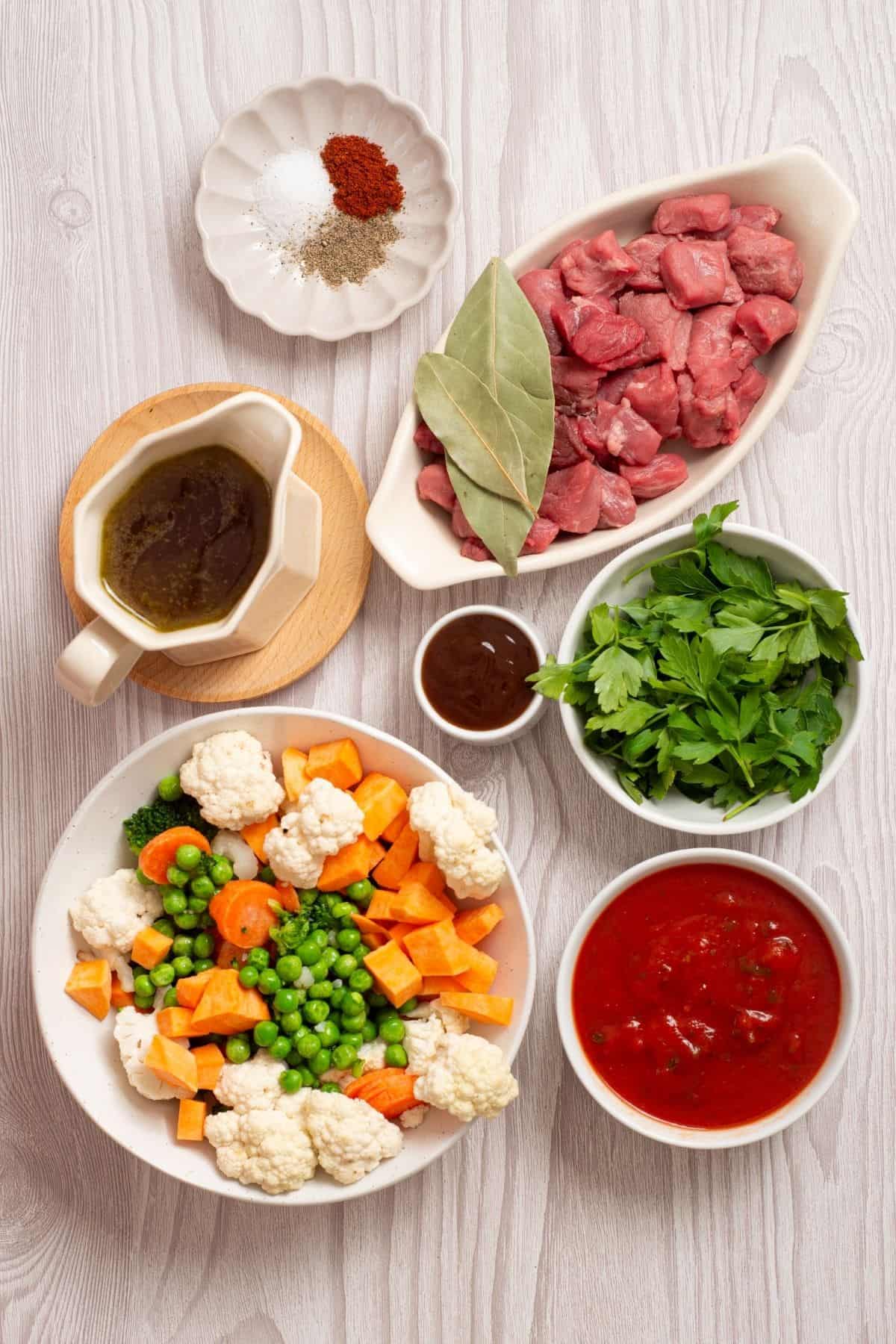 Ingredients for a meal arranged on a wooden surface, including cubed beef, chopped vegetables, parsley, tomato sauce, seasonings, and a gravy boat.