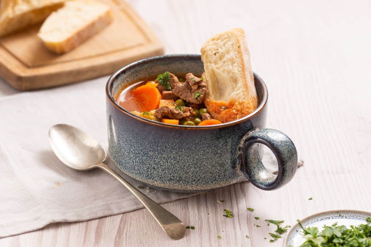 A ceramic bowl of beef stew with carrots, peas, and a piece of bread, served with a spoon.