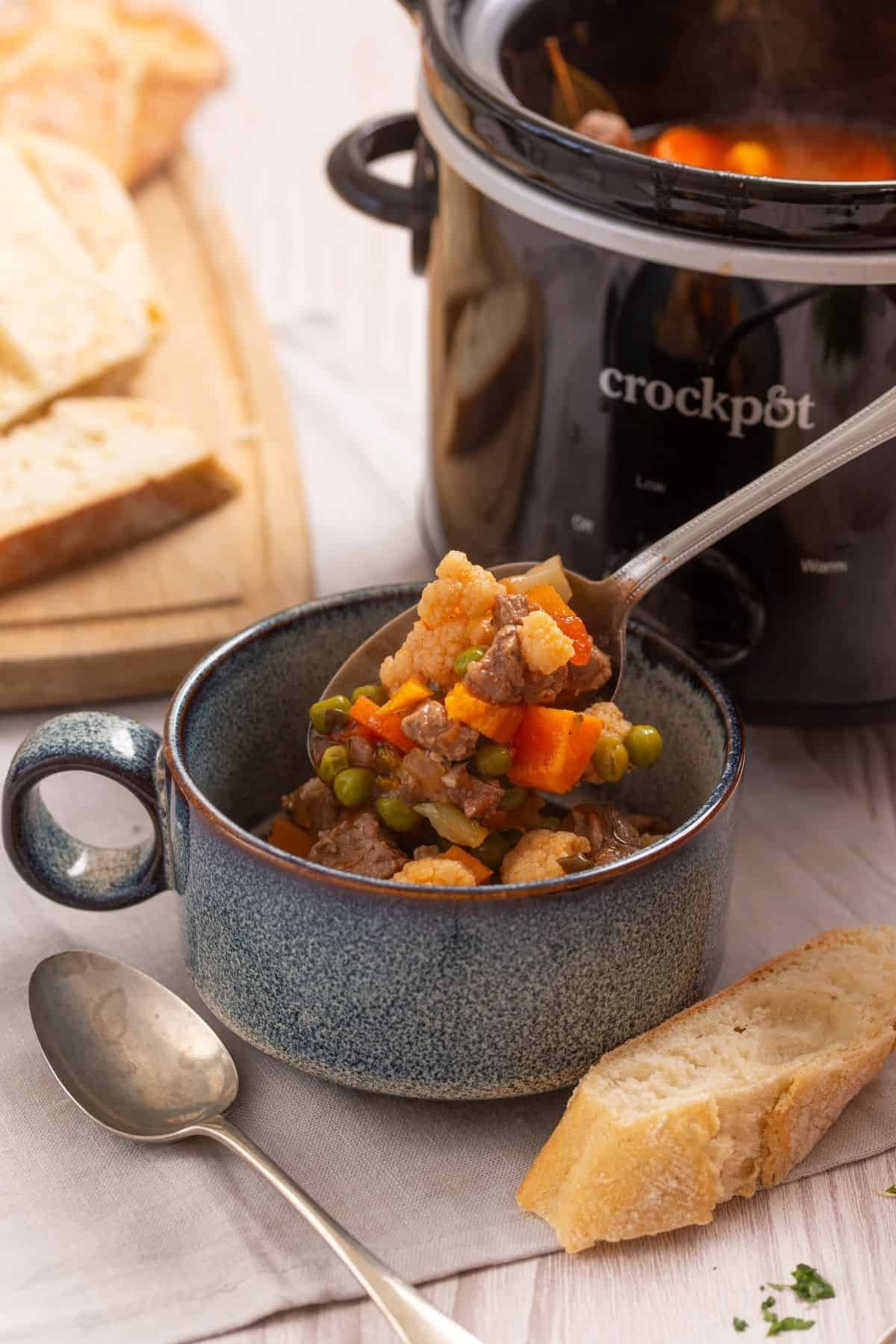 A spoonful of vegetable and beef stew held over a ceramic bowl, with a Crock-Pot and slices of bread nearby.