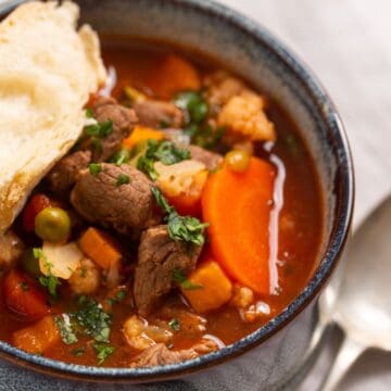 A bowl of beef stew with carrots, peas, and cauliflower, served with a piece of bread.
