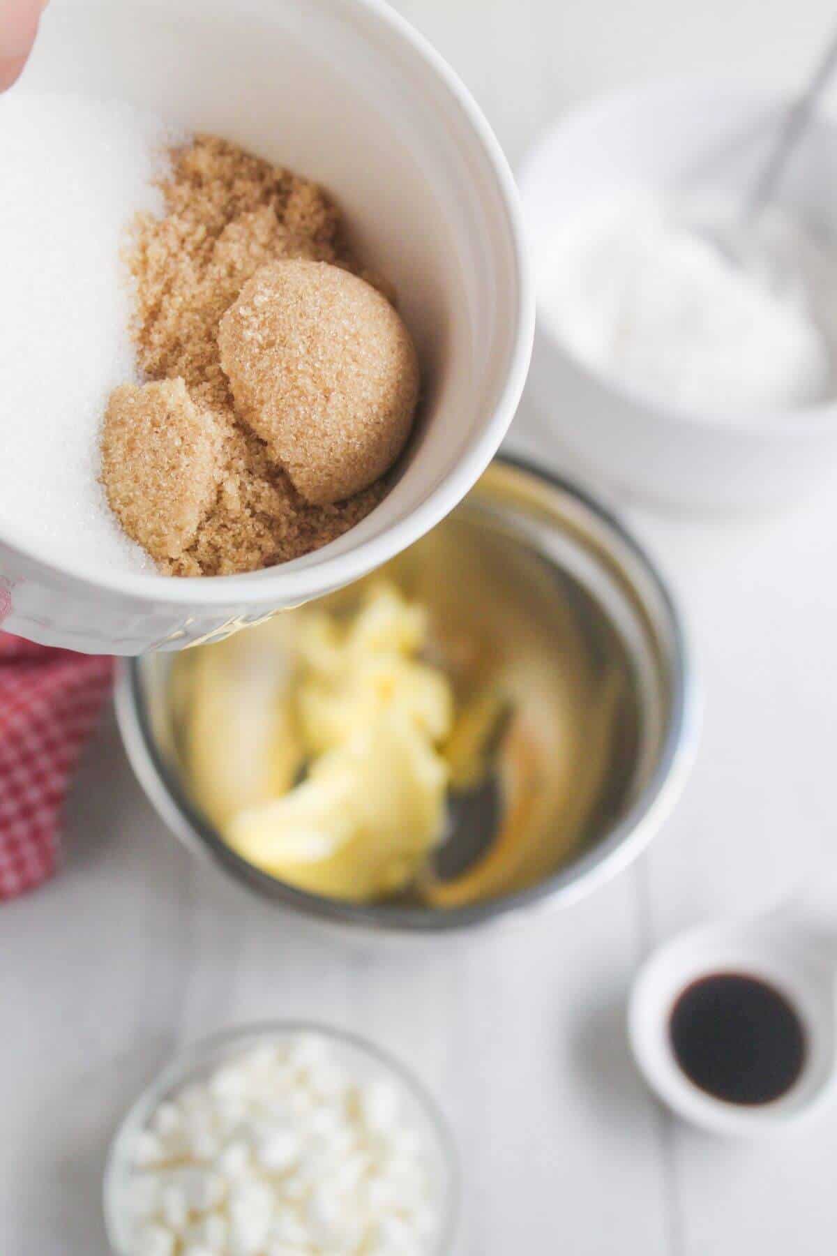 A hand holds a bowl containing white and brown sugar over a mixing bowl with butter.