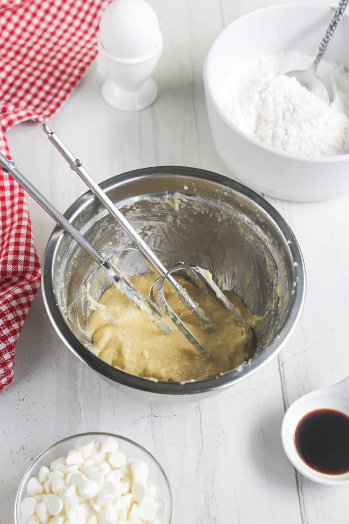 A metal mixing bowl containing dough with two electric beaters, surrounded by a bowl of flour, a small bowl of dark liquid, white chocolate chips, and an egg.