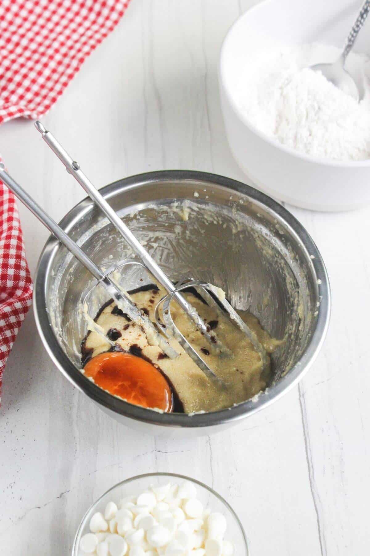 A hand mixer inside a stainless steel bowl with cookie dough, an egg, and vanilla extract, next to a bowl of flour and a bowl of white chocolate chips.