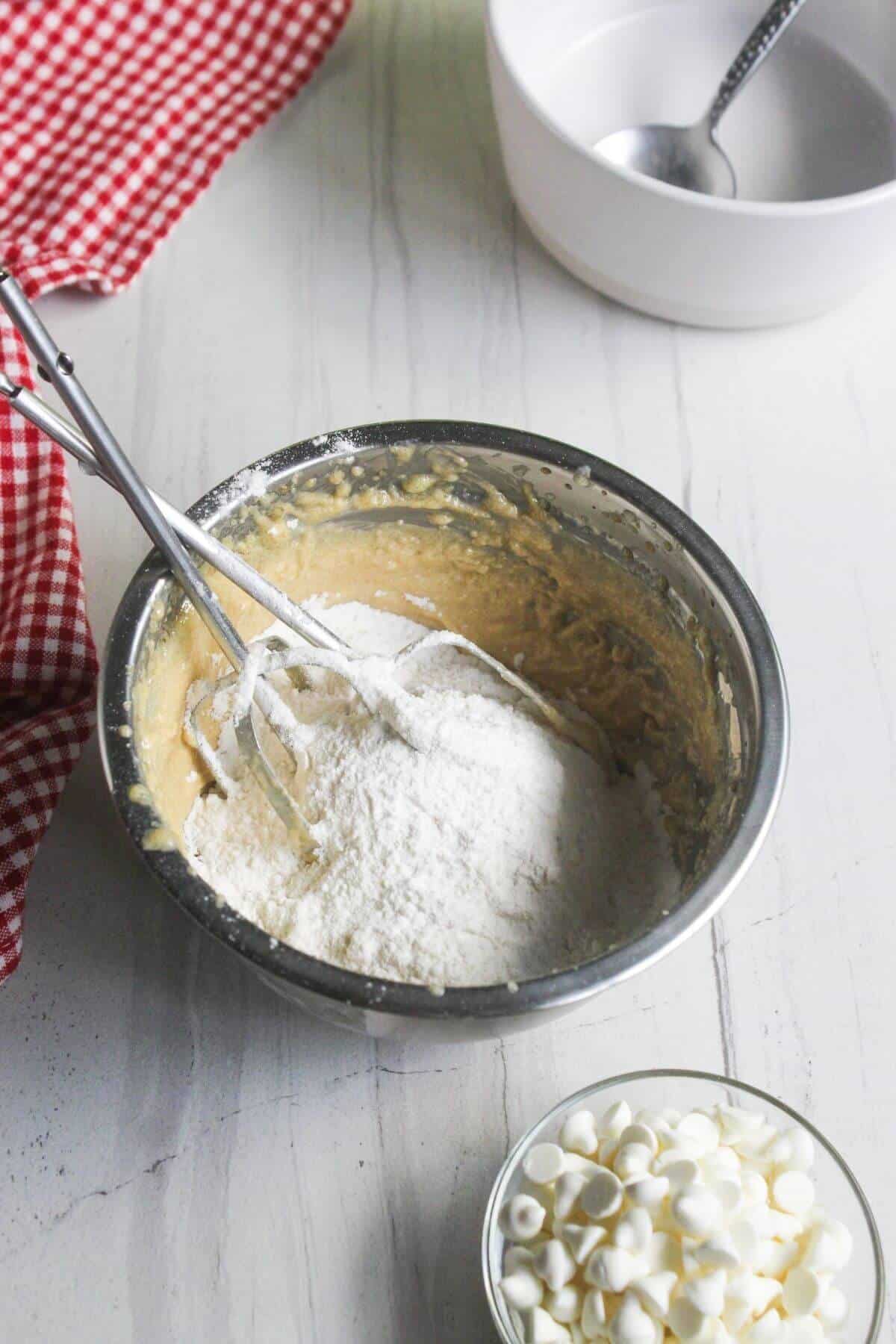 A metal mixing bowl containing cake batter and flour, with electric beaters resting inside, next to a small bowl of white chocolate chips.