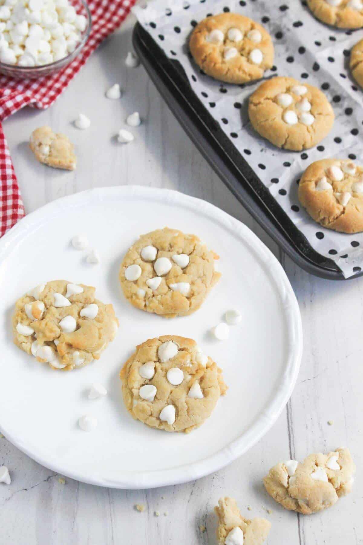 Three white chocolate chip cookies on a white plate, with more cookies on a baking sheet nearby.