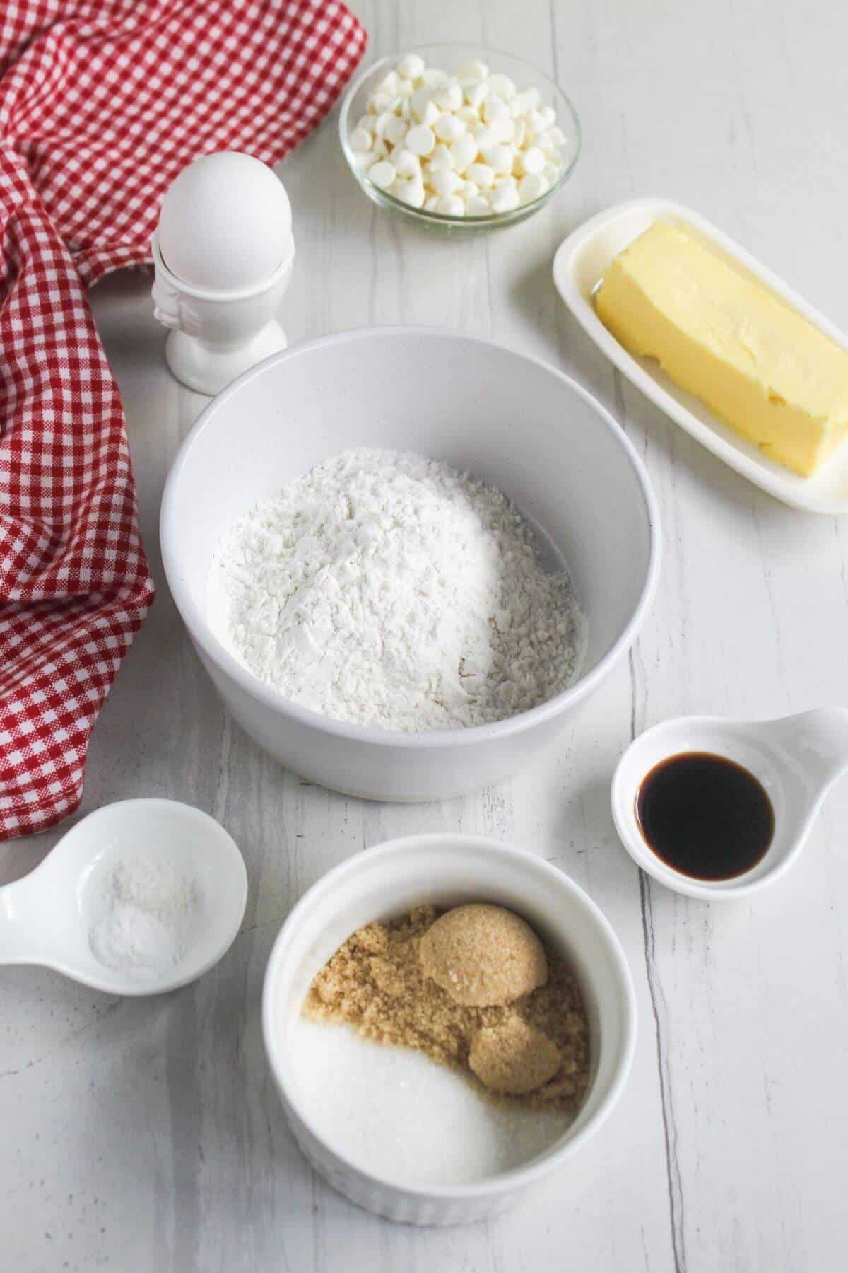 Baking ingredients including a bowl of flour, brown and white sugar, white chocolate chips, butter, an egg, vanilla extract, and salt.