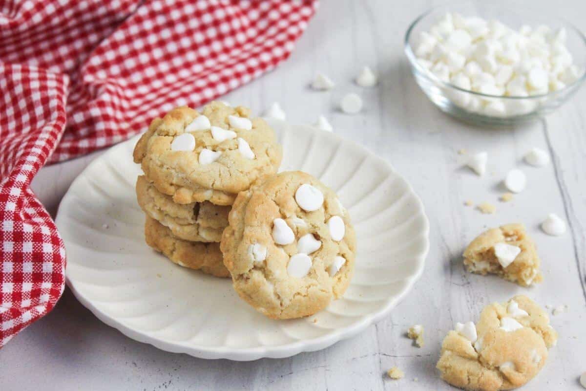 A stack of white chocolate chip cookies on a fluted white plate, with a bowl of white chocolate chips nearby.