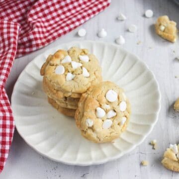 A stack of white chocolate chip cookies on a scalloped plate next to a red and white checkered napkin.