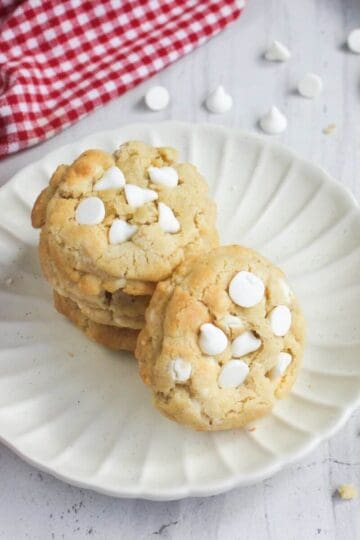 A stack of white chocolate chip cookies on a scalloped plate next to a red and white checkered napkin.